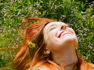 Woman with red hair smiling, lying in a field of green grass and purple flowers. Bright, sunny day, evoking joy and relaxation.