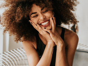 Woman with curly hair smiling joyfully, hands on cheeks. She wears a black top, sitting indoors on a striped chair. Bright and cheerful mood.