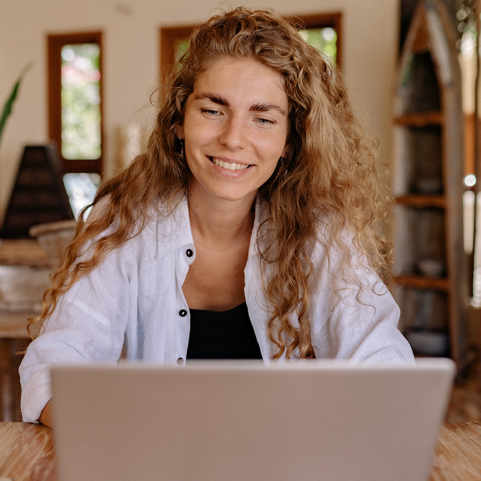 Woman with curly hair smiling at a laptop in a cozy room with wooden decor and large windows. She wears a white shirt, conveying a relaxed mood.