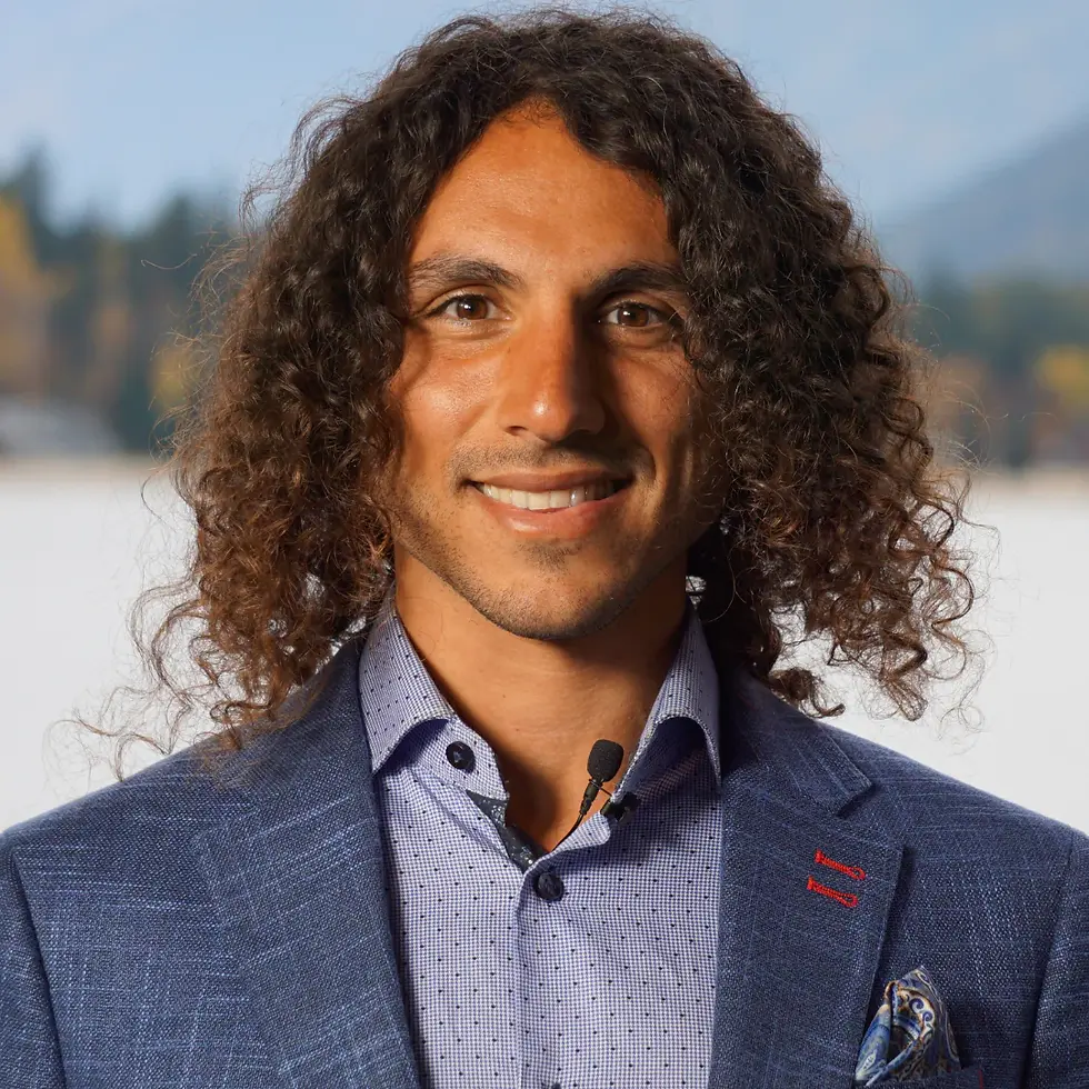 Curly-haired person smiling, wearing a blue blazer and shirt with a microphone. Blurry scenic background suggests an outdoor setting.