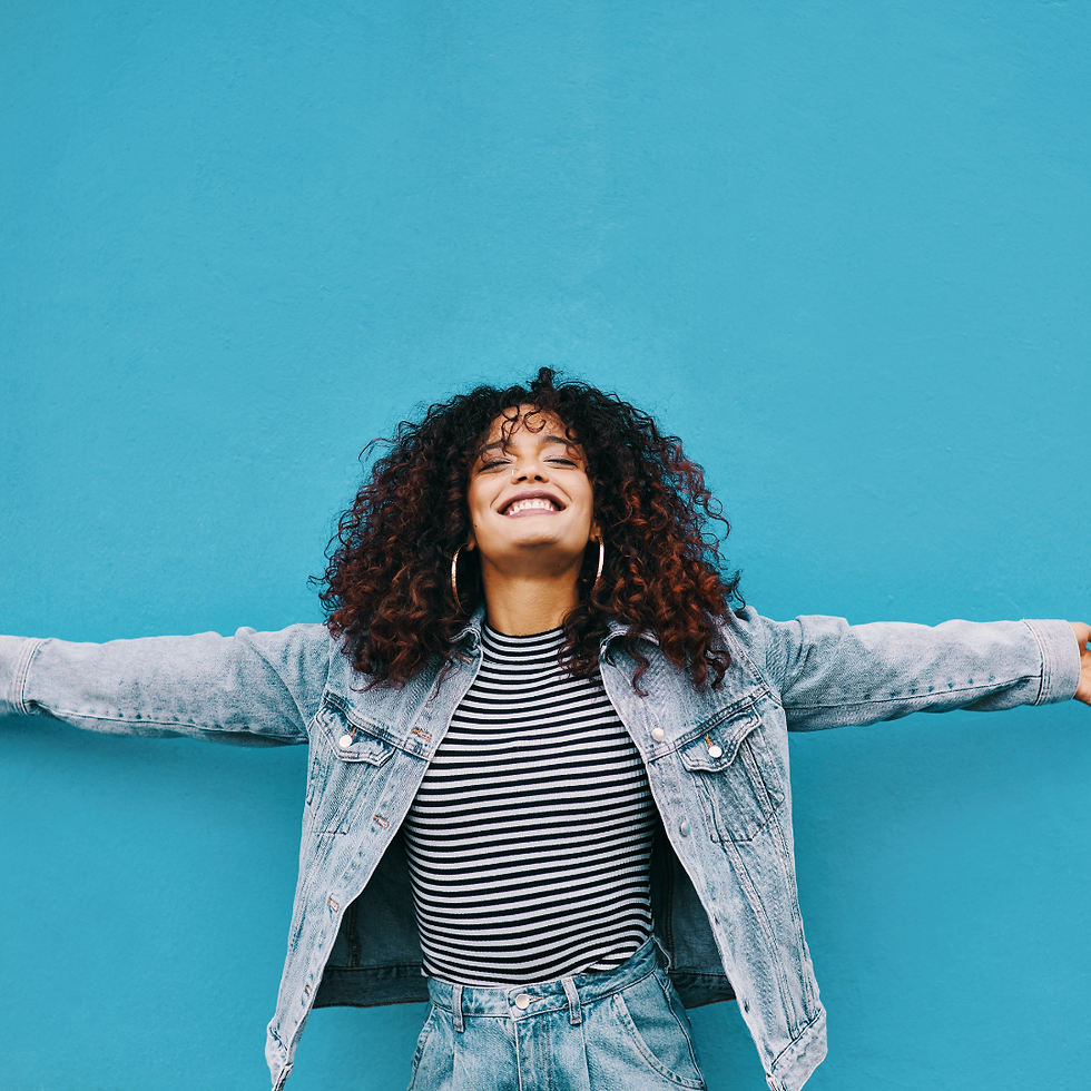 Woman with curly hair smiles, arms outstretched against a bright blue wall. She wears a striped shirt and denim jacket, exuding joy.