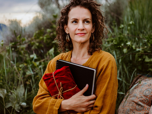 Curly-haired woman in a mustard dress holds a red-cloth-wrapped notebook. She's outdoors with greenery, looking content.
