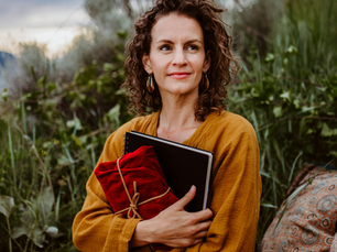 Curly-haired woman in a mustard dress holds a red-cloth-wrapped notebook. She's outdoors with greenery, looking content.