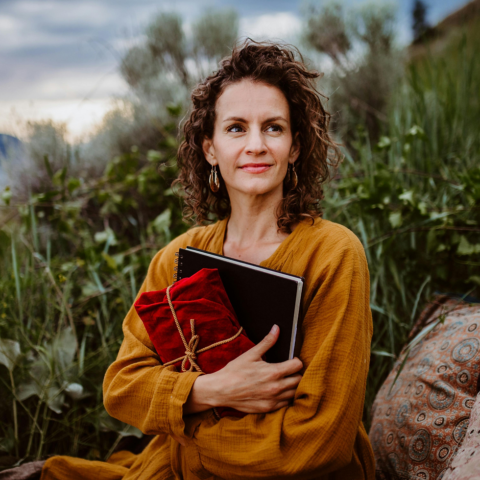 Curly-haired woman in a mustard dress holds a red-cloth-wrapped notebook. She's outdoors with greenery, looking content.