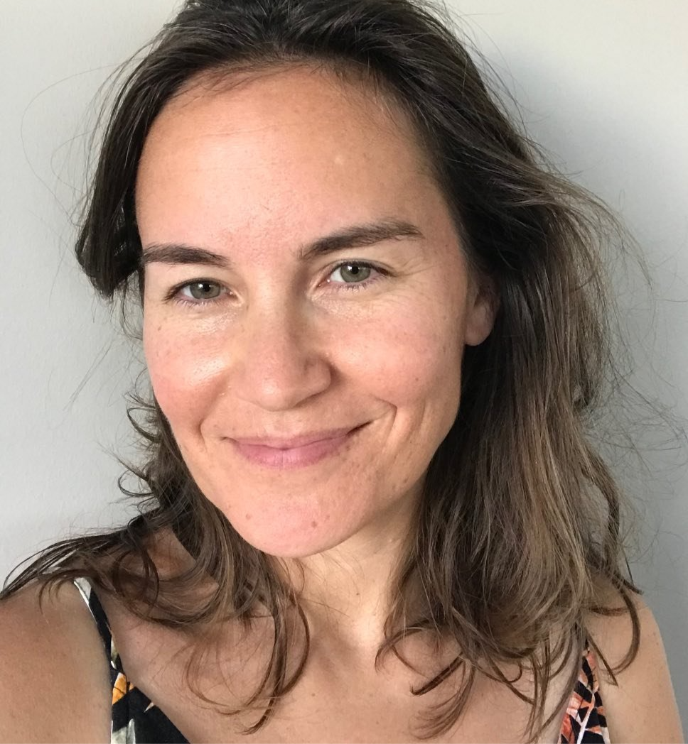 A woman with brown hair smiles softly against a plain background. She wears a patterned dress with earthy tones, exuding a relaxed mood.