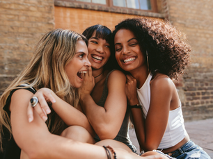 Three women sit closely, laughing joyfully against a brick wall. The scene is warm and casual, capturing a moment of friendship and happiness.