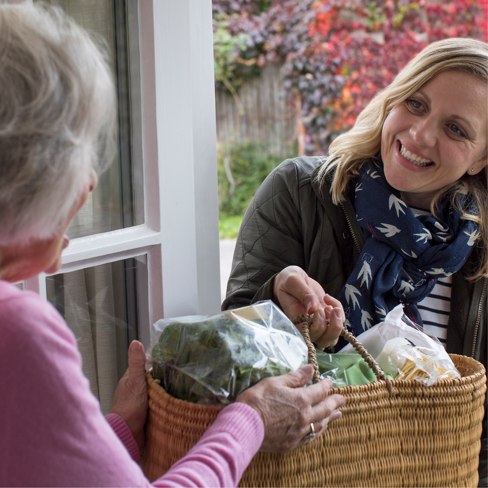 Woman delivering groceries to an elderly woman at a door. She's smiling, wearing a scarf with bird patterns. Autumn foliage in background.
