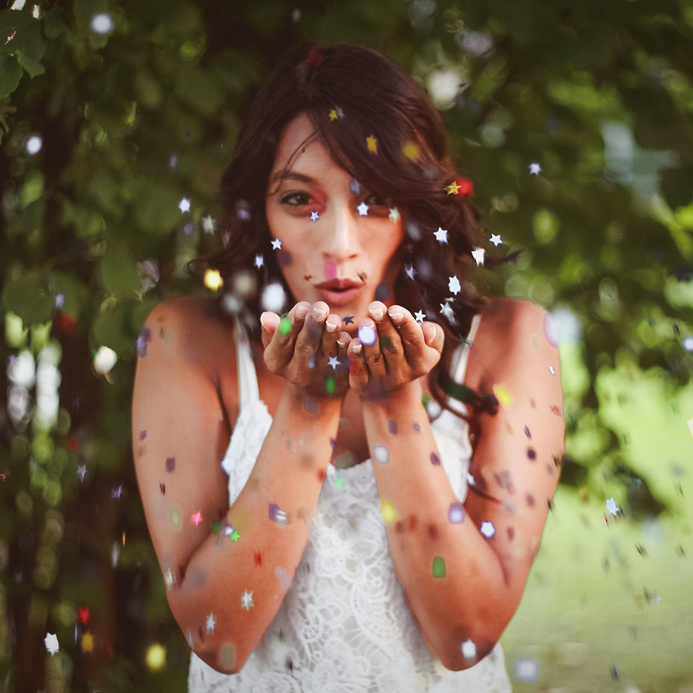 Woman in white dress blows colorful confetti from hands, surrounded by green foliage. Playful and joyful mood.
