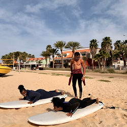Leandro, instructor of the island kite school, is teaching surfing with to local girls. All three are wearing a black wetsuit. He is standing and the girls laying on the surfboards at the Beach of Santa Maria. 