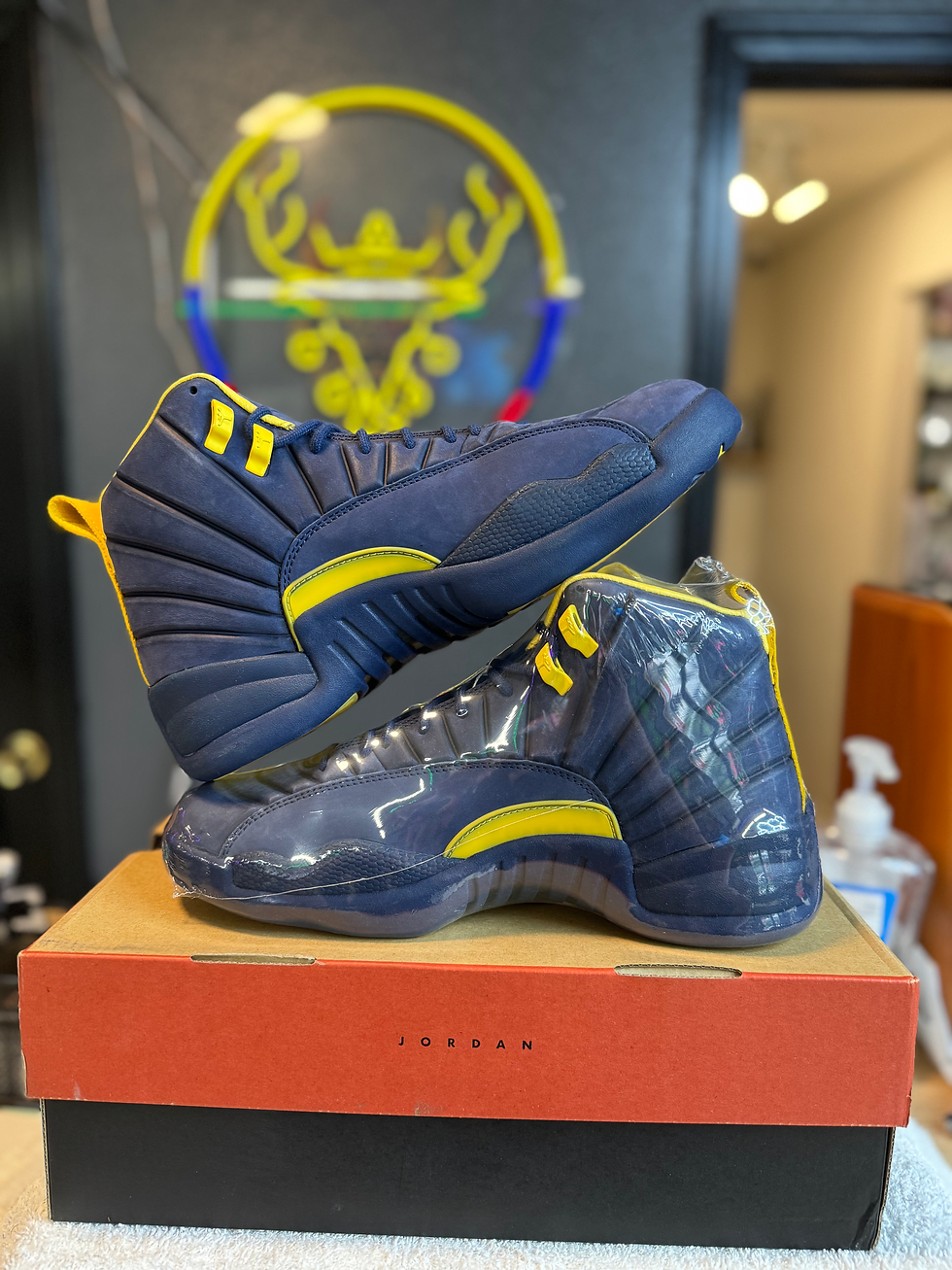 Thumbnail: Two JORDAN 12 PSNY MICHIGAN PE sneakers, navy and yellow, on shoebox.