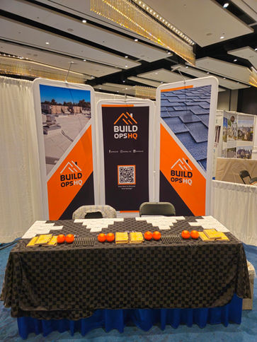 Stand de Build Ops HQ en una feria comercial con pancartas de la marca, folletos, bolígrafos y pelotas antiestrés naranjas organizados sobre un mantel negro, dentro de un salón de exhibición.