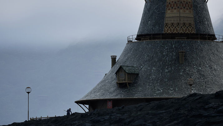 a large, conical building with intricate designs on its upper sections. Its shingled exterior and wooden elements give it a rustic, medieval appearance. The setting is moody, with misty hills or mountains in the background. A lone figure with a stick or staff is seen near the base of the structure, adding a sense of scale and solitude. The scene evokes a mysterious, cinematic atmosphere.