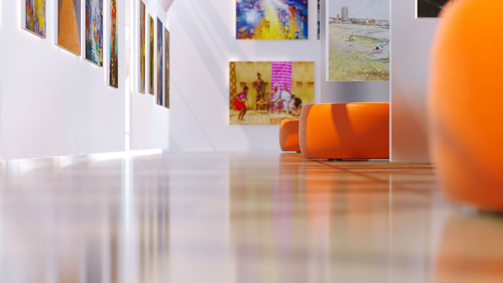 Side view of an art gallery with a row of colorful paintings on white walls, glossy wooden floors, and orange cushioned seating.