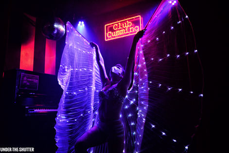 A dark photo of a stage at Club Cumming, NYC of a woman wearing a silver mask and light-up wings, which are spread out.