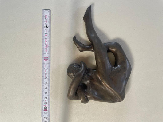 A small bronze sculpture with a dark greenish-brown patina and a smooth, slightly glossy finish, depicting a voluptuous female figure lying on her back, captured from a direct overhead view. Her upper torso and head are curled deeply forward, with her thick arms wrapped tightly around her chest and bent head. Her long, full legs are dynamically thrust upward into the air; one leg is extended straight and nearly vertical, while the other is bent at the knee and angled outward. To the left of the sculpture, a white and yellow folding ruler is placed vertically, showing centimeter markings up to approximately 20 cm. The entire setup is photographed against a plain, light neutral background.