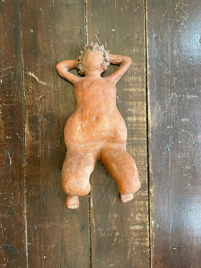 Top-down view of a clay sculpture in a warm terracotta-orange hue, depicting a female figure reclining on her back on a dark wooden plank floor. The figure is styled with voluptuous, organic curves, her arms folded behind her head, and blurred facial features. Her lower body and thighs are prominently wide, with knees slightly raised and bent upward, ending in small feet resting on the floor.
