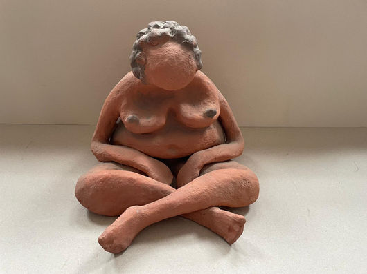 A terracotta clay sculpture depicting a voluptuous female figure sitting cross-legged. The figure's head is bowed deeply forward, with a smooth, faceless head topped by dark, textured, curly hair. Her relatively long, slender arms extend downward with her hands resting gently on her thighs. The sculpture is captured from a straight-on, frontal view against a plain, neutral background.