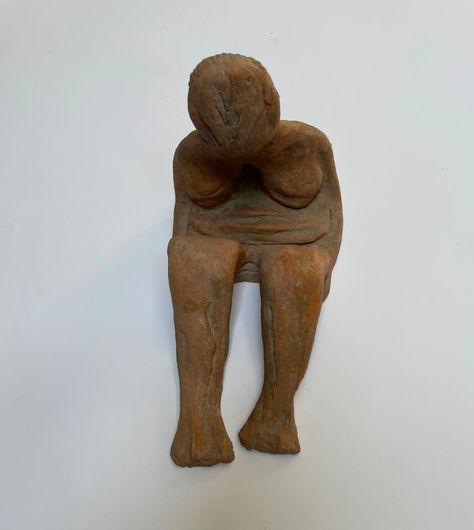 Earthy terracotta clay sculpture of a seated nude figure with a bowed head and a raw, natural texture.