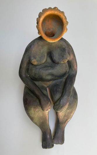 A dark bluish clay sculpture of a seated woman resting her hands on her legs, featuring an open round cavity at the top of her head.