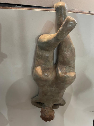 A bronze sculpture with a greenish-brown patina and a textured matte finish, depicting a voluptuous female figure hanging upside down, captured from a straight-on eye-level view. The figure hangs vertically, suspended by a single straight leg hooked over the edge of a top surface, while her other leg is bent at the knee and crossed forward in a dynamic pose. The lower body features prominent, rounded hips and buttocks, tapering downward toward a slender waist and shoulders. Her arms are folded behind her downward-facing head, which is topped with textured, rusty-brown curly hair. The sculpture hangs against a light, neutral background, casting a soft shadow to its left.