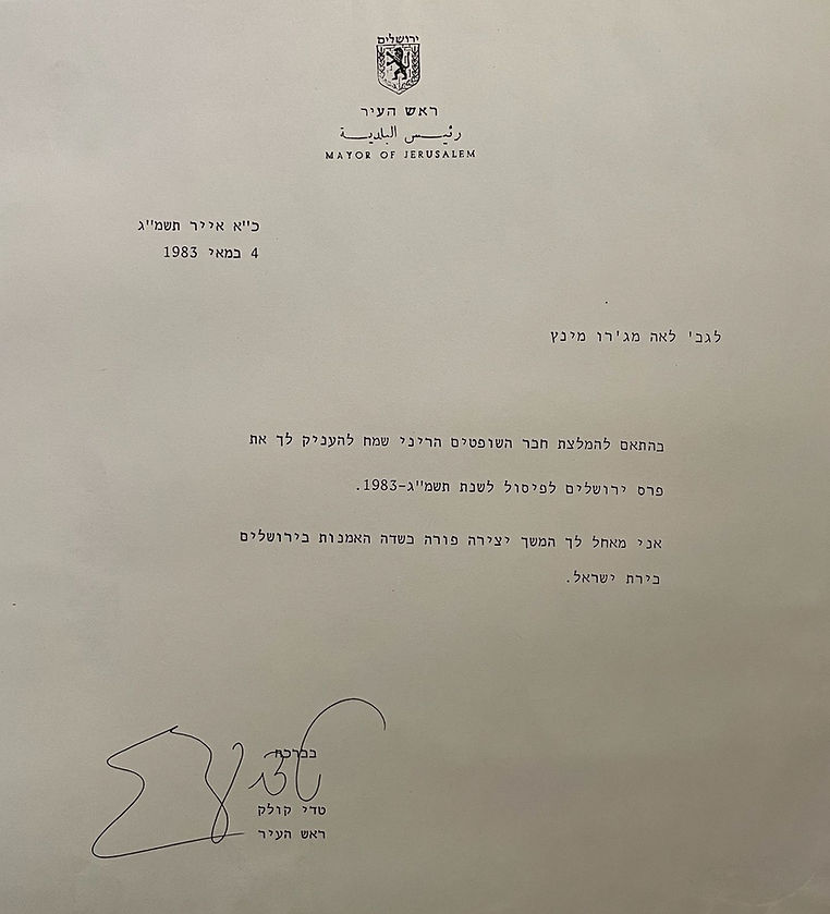 A documentary color photograph from an official ceremony in 1983. In the center, the Mayor of Jerusalem, Teddy Kollek, dressed in a dark suit and light shirt, is smiling as he hands the Jerusalem Prize certificate to the artist Lea Majaro-Mintz. Lea, looking back at him with a smile, wears a light-colored dress with a subtle floral pattern and a necklace. The event takes place indoors, with other attendees visible in the background watching the presentation.