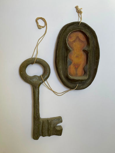 A close-up, frontal photograph of a complex ceramic sculpture. On the right, a large, heavy, dark bronze-like lock in an oval shape. Inside the deep central cavity of the lock, bathed in warm, luminous shades of gold and copper, is a sculpted silhouette of a full-figured woman. To the left, a corresponding oversized, rustic, dark key is connected to the lock by a thin natural fiber rope.