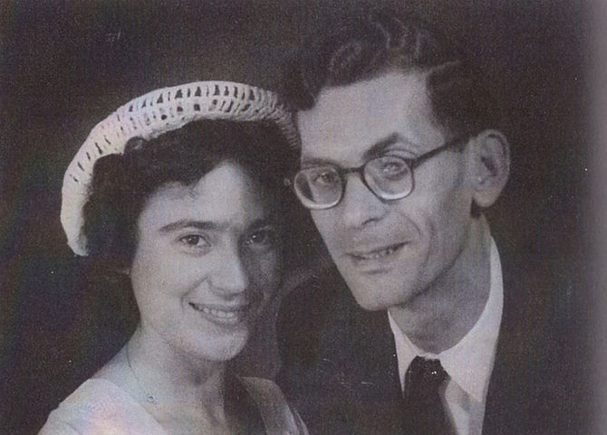 A historic black and white wedding photograph of Lea and Isaac on their wedding day in 1951. Lea, on the left, is smiling and wearing an elegant dark long-sleeved wedding dress with a deep V-neckline and a light flower brooch on her lapel, while holding a small bouquet of light flowers. Isaac, on the right, is smiling and dressed in a dark suit, a white shirt, and a dark necktie. The background is a plain, light-colored wall.