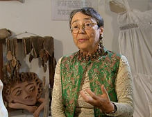 Lea Majaro-Mintz in a still image from a 2003 television interview. Lea is captured inside the historic Rokach House in Neve Tzedek. She wears glasses, a statement necklace, and a green patterned vest over a light-colored long-sleeve top, captured mid-gesture during her conversation. The background showcases her artwork and sculptures, including a prominent ceramic face sculpture looking sideways to her left, and a white garment hanging to her right.