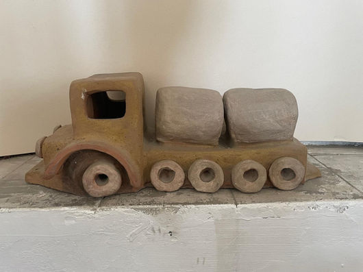 Profile view of a clay sculpture depicting a large cargo truck, resting on a light-colored surface against a plain white wall. The truck's cabin and chassis are crafted from a textured mustard-brown clay, featuring a hollow square window and a large front wheel. The rear section displays four smaller wheels lined up in a row, carrying two rounded, blocky cargo containers or cylinders made of a smooth gray clay.