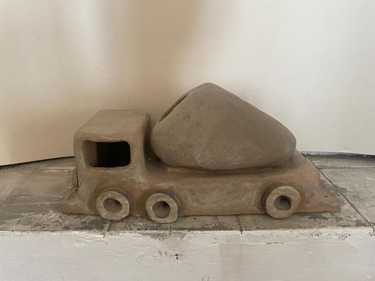 Profile view of a clay sculpture depicting a cement mixer truck, resting on a light-colored ledge against a plain white wall. The piece is crafted from raw, natural grayish-brown clay. The truck features a blocky driver's cabin with a hollow window cutout, three prominent round clay wheels, and a large, teardrop-shaped mixing drum mounted on the back. The overall design has a simple, naive, and rustic quality.