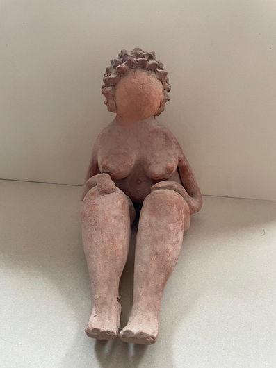 Front view of a clay sculpture in a natural terracotta hue, depicting a nude female figure sitting on the floor and leaning back against a white wall. The figure is styled with simple, rounded contours, featuring short textured hair and smooth, featureless facial details. Her slender arms rest on her voluptuous thighs, and her legs extend straight forward in a parallel position.