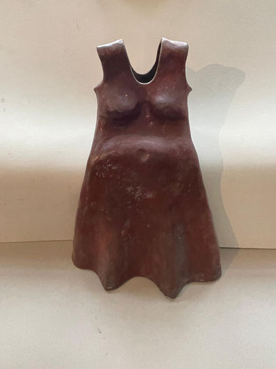 A dark chocolate-brown bronze sculpture with a smooth, slightly glossy finish, shaped in the form of a sleeveless women's dress with wide straps and a round neckline. The dress is hollow at the top opening and takes on the contoured shape of a voluptuous female torso, with an emphasized bust and a prominent, rounded belly. The lower section of the dress flares outward and terminates in wavy, undulating edges that mimic the drape of fabric. The sculpture is captured from a straight-on, frontal view against a plain, light background, casting a distinct shadow on the wall to its right.