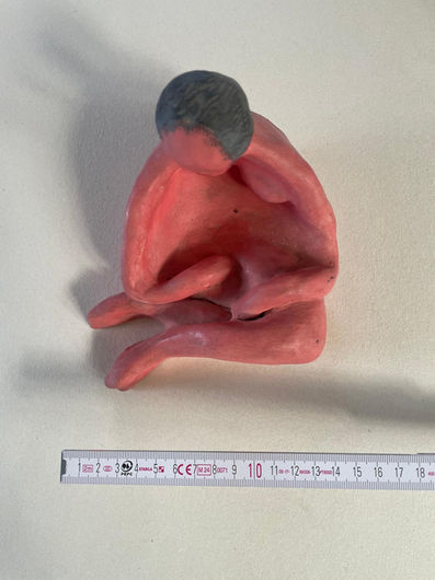 A small clay sculpture depicting a seated human figure, captured from an overhead (bird's-eye) perspective against a plain, light background. The sculpture features a distinct painted color scheme: the entire body is painted a vibrant bubblegum pink, while the top of the head is painted a smooth dark gray, suggesting short hair. The figure is bent forward, with long, rounded arms extending downward to encircle a hollowed center torso, and hands resting on the bent legs. At the bottom of the frame, a white folding ruler is laid horizontally, displaying centimeter markings from 1 to 16 cm to show the scale of the artwork.