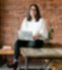 Casey Hanisko, long dark hair, white skin, glasses. Wearing a white sweater and holding a laptop. Brick wall and plants in the background.