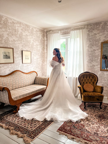 Brud som står med ryggen mot kameran framför ett fönster och tittar sig över axeln