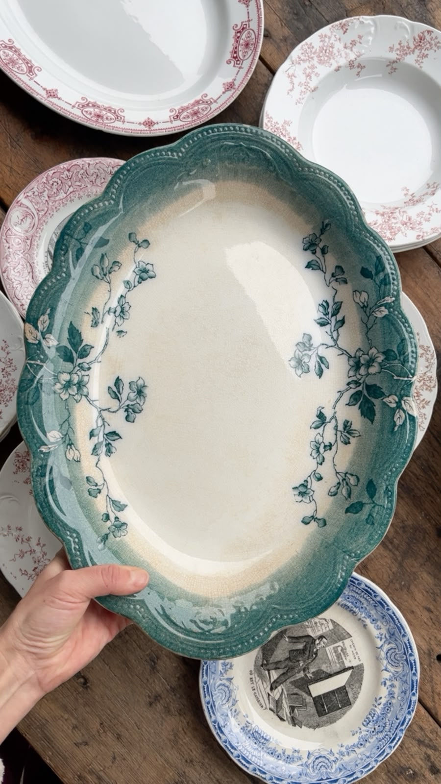 Mano sostiene plato ovalado verde con flores; alrededor hay platos blancos y azules vintage sobre una mesa de madera.