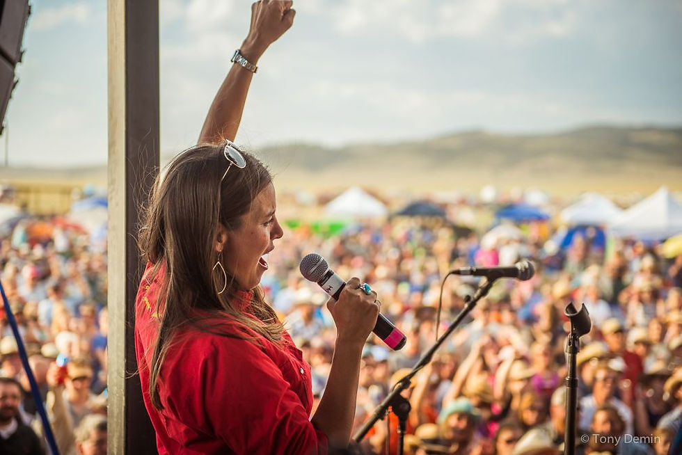 Woman in a red shirt sings into a microphone with one arm raised, facing a cheering crowd at Red Ants Pants Music Festival