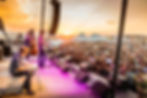Corb Lund plays on an outdoor stage at sunset, with a huge crowd, white tents, and glowing orange-pink sky at Red Ants Pants Music Festival.