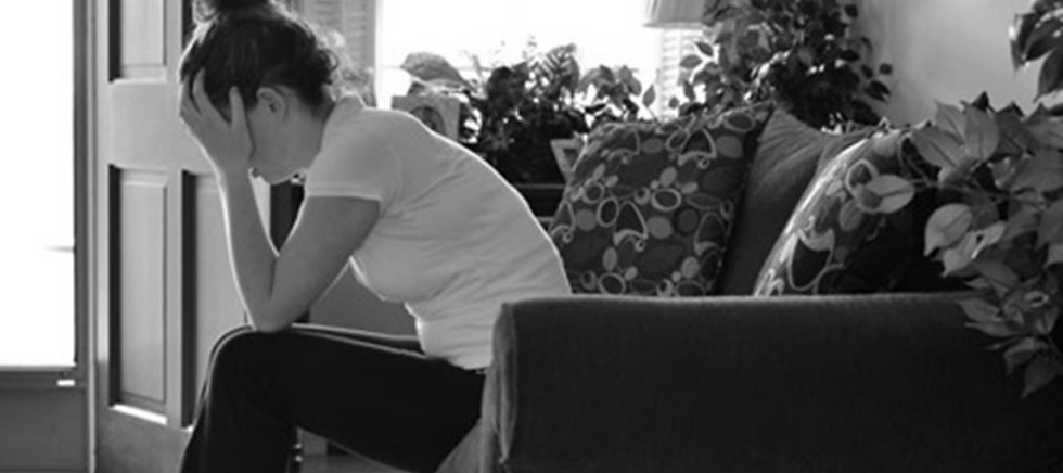 Black-and-white photo of a woman sitting on a couch, head in hands, looking distressed in a plant-filled living room.