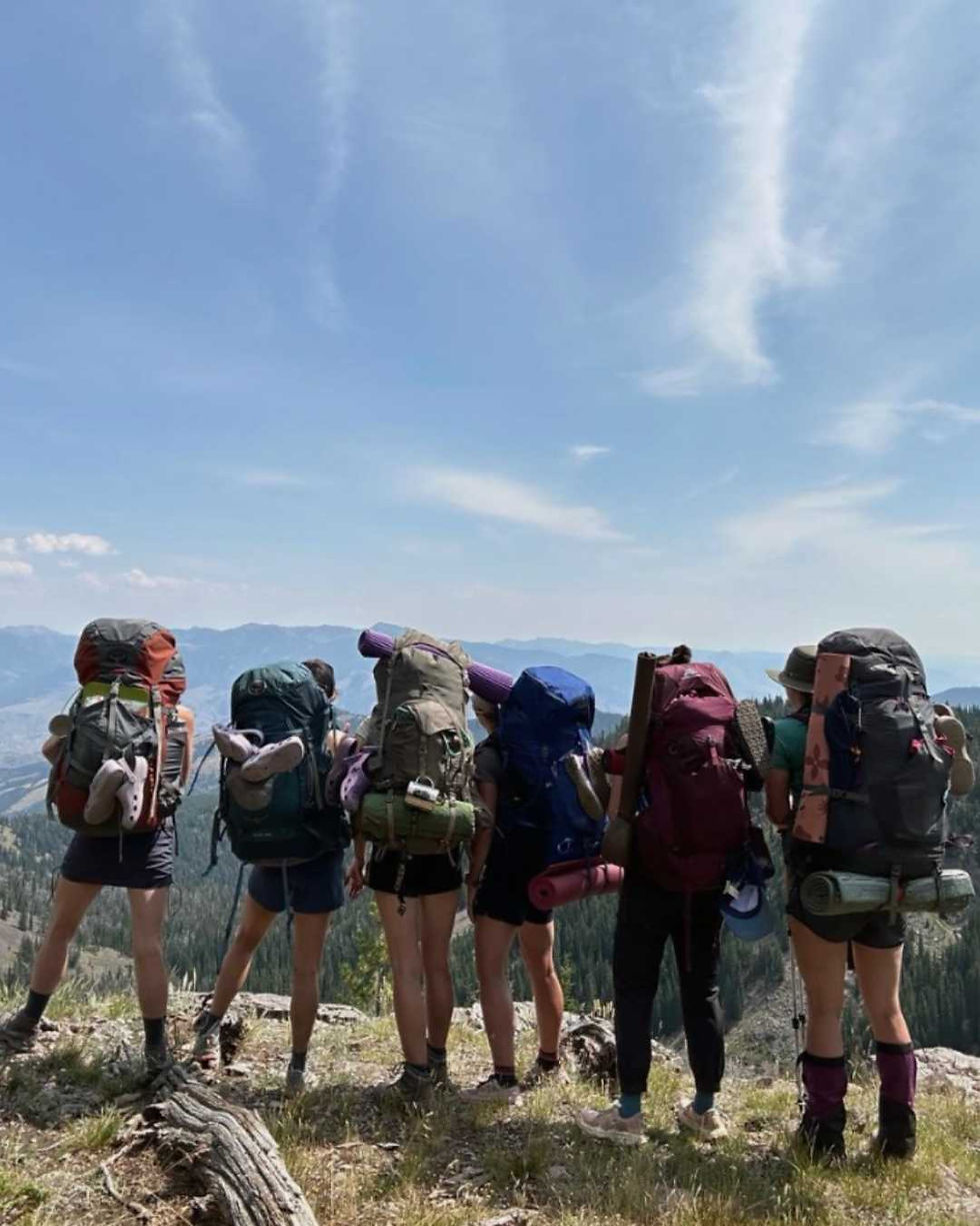 A group of women with large hiking backpacks stand together at a mountain summit, looking out over a sweeping panoramic view