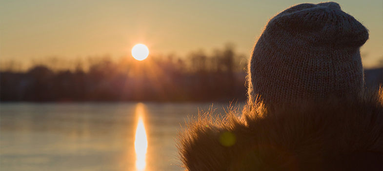 Person in a knit hat and fur collar смотрing a golden sunset over a calm lake, with trees silhouetted behind