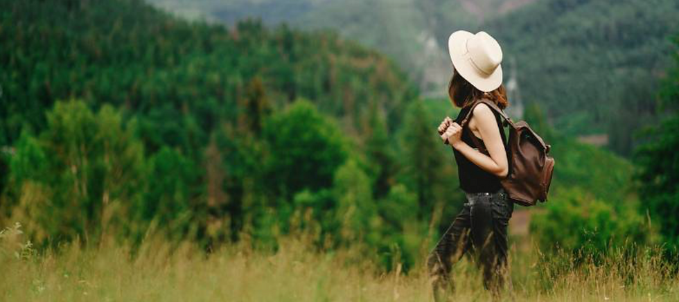 Woman hiker in a white hat and backpack stands in tall grass, gazing at green forested mountains.