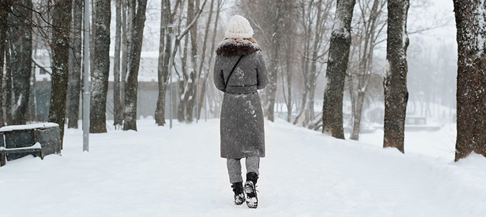 Woman in gray coat and white knit hat walks away through a snowy, tree-lined park in falling snow.