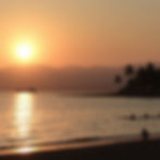 Sunset over a calm sea with a distant boat, swimmers, and silhouetted palm trees on the shore. Sky is orange, creating a serene mood.