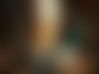 Elderly woman in green sari prepares food by open fire in a rustic kitchen. Clay pots and wood logs are visible, creating a warm, earthy ambiance.