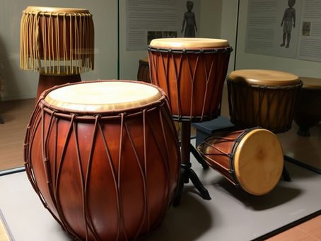 Wooden hand drums with rope lacing on display in a museum setting. Informational text panels in the background. Warm lighting.
