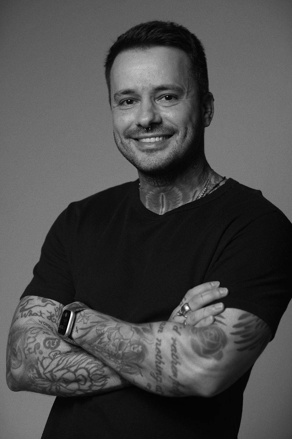 Retrato em preto e branco de tatuador sorrindo, com tatuagens visíveis no braço, Kreyner La Scala Tattoo Ateliê