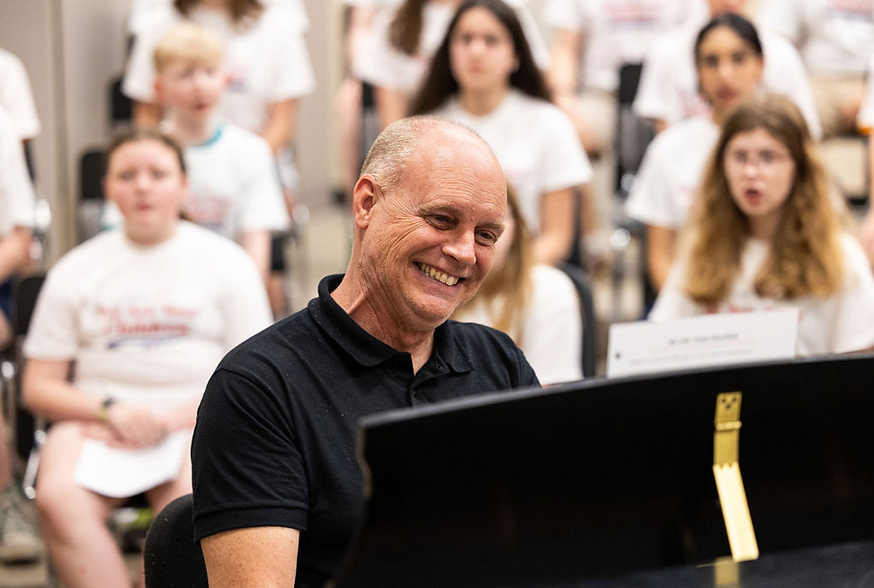 A smiling man plays piano for we are your children choir.