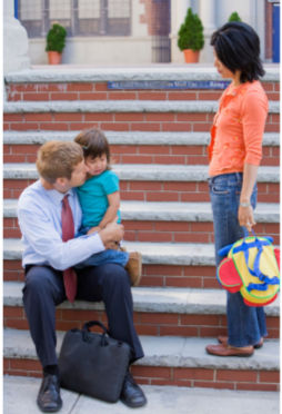 A father in work clothes sits on school stairs holding a distressed young girl cheek to cheek while a woman stands quietly a few feet away, backpack at her side, watching