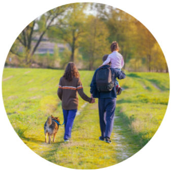 A family walking together on a grassy path — a father carrying his young daughter and a backpack while a mother holds his hand and walks the dog, representing the everyday road of family life.